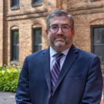 a photo of Paul Avey, wearing a suit, standing in front of a collegiate-looking building